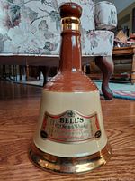Full view of the porcelain whiskey decanter standing upright on wood floor with floral sofa background. Shows bell shape, color tones, and stopper.