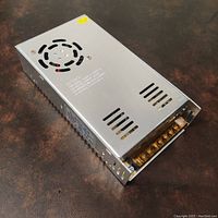 Top view of the silver metal enclosed rectangular power supply unit showing cooling fan, ventilation slots and printed specs.
