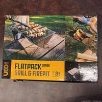Box front showing product usage with grill cooking skewers and firepit in use in a camping setting.