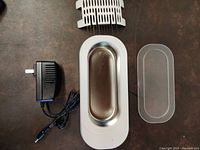 Top-down view of ultrasonic cleaner and included power adapter, plastic cover, and basket insert on a brown surface.