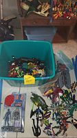 Large blue bin filled with assorted loose action figures, mostly colorful characters from X-Men, with many posed differently and some lying down. A labeled sticky note marked 'Action Figures' is visible. Around the bin are plastic bags containing metal planes and other toys. Shelf in background holding more toys is partially visible.