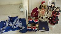 Wide shot showing blue Greek jacket with white embroidery, Greek figurines, shoes, decorative hat, and a book with emblem and year 2015 on cover.