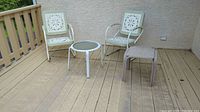 Two metal chairs and round metal side table on wooden deck, plus unrelated plastic stool