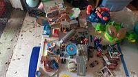 Overhead photo showing multiple Micro Machines playsets including gray roadway with runway markings, helipad, and rocky landing pad. Various small cars and Bucky O'Hare figures and toys are placed around the playsets with a small stack of baseball cards.