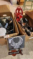 Two boxes filled with assorted motorcycle parts including a red multi-trumpet horn, partial handlebar, and various hardware components with Harley-Davidson patches and a framed print positioned in front of the boxes.