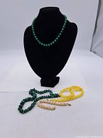 Green beaded necklace displayed on black jewelry bust with bracelets and necklace in front.