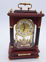 Front and partial side view showing clock face with Roman numerals and pendulum of gold-tone spheres inside wooden case with glass panels