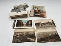 Overhead view of a large assortment of vintage and antique photographic postcards laid on a white surface, showing various black and white and hand-colored images.