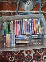 Top-down view of a plastic bin filled with assorted VHS tapes in cases shown on a patterned rug. Titles like The Sound of Music, Pinocchio, and Finding Nemo are visible.