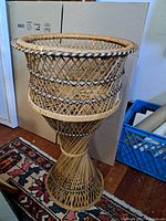 Full view of tall vintage cane jardiniere stand with lattice weaving, showing basket top, tapering center, and flared base on floor with carpet.