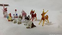 Front view of 9 small porcelain animal figurines including roosters, ducks, dogs, and deer all positioned on a white surface.