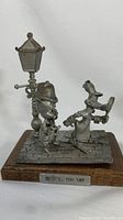 Front and side view of the metal figurine showing Daffy Duck with magnifying glass and Porky Pig in detective outfits on cobblestone base with lamp post attached to wooden stand.