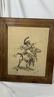 Framed art on rawhide showing Native American warrior on horseback, image signed by Frederick Remington. Frame is wooden with visible wear; rawhide has stains and discoloration.