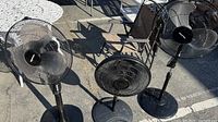 Three black adjustable oscillating pedestal fans of varying sizes positioned outdoors on the ground under daylight. Fan heads measure approximately 16, 14, and 14 inches in diameter respectively, with circular protective metal grilles and plastic fan blades.