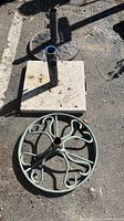 Photo showing three umbrella stands on pavement: a green round stand with ornamental swirls at the front, a heavy square stone base behind it, and a gray round base further back