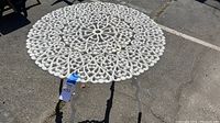 Close-up front angled photo showing the intricate white wrought iron scrollwork on the round tabletop with the umbrella hole in the center, highlighting the design and condition.