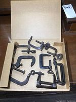 Photo showing all 8 small cast metal C clamps in a cardboard box on a wooden surface, arranged loosely to display variety in size and shape.