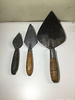 Photo showing all three trowels lined up by size from smallest to largest, displaying metal blades and wooden handles with aged patina.