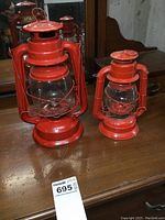 Photo showing both red kerosene lanterns together on wooden surface with mirrored background.