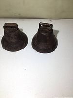 Pair of old metal farm bells placed side by side, showing general size and shape with aged surface and rust. Both bells appear similar in condition and design.