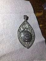 Front photo showing full pendant with detailed filigree and central medallion design on a neutral fabric background.