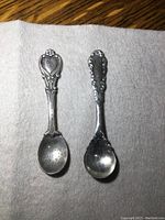 Two sterling silver salt spoons shown on a white cloth, displaying the full spoons with decorative handles and small bowls with some tarnish.