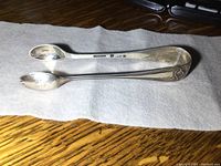 Paired vintage sterling silver sugar tongs placed on white cloth on wooden surface showing top side with hallmarks and decorative design.