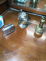 Three vintage free standing metal lighters on wooden surface: two knight helmet shaped with detailed engravings, one brass-colored pump-style lighter.