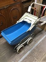Side angled view of the vintage metal doll stroller showing blue plastic body, white frame, canopy, and wheels with spring suspension.