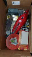 Top view of a box with assorted hardware items including Dirt Devil vacuum, drill bits, screws, funnel, and a hand caulking tool.
