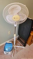 Full view showing standing fan in front of two black folding tables, and humidifier on the floor near the fan stand.
