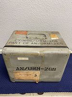Closed metal carrying case with label AN/URM-26B on lid and military data tags.