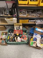 Overview of various tools, hand tools in green container, assorted hardware items, and bulbs spread in boxes on floor