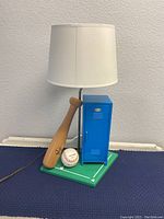 Full view of the baseball locker lamp showing the bat, baseball, blue locker, green base, and white lamp shade against a white wall.