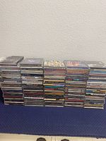Photo showing large stacks of 136 assorted music CDs in jewel cases, arranged horizontally in multiple piles on a flat surface.