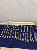 Array of over 20 souvenir spoons laid out on a dark fabric background, showing different sizes, finishes and decorative handle tops including enamel emblems and city names.