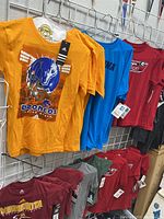 Lot of assorted kids collegiate T-shirts hanging on a rack showing multiple colors including orange, blue, maroon, and gray with team logos visible