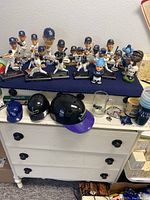 Overview of lot showing Tampa Bay Rays bobbleheads arranged on countertop, two helmets, mugs and glass tankards below on dresser