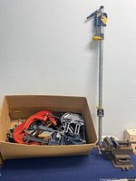 Full view of lot showing a large bar clamp with yellow and black quick-release clamp, a box of various red and metal C-clamps, and a heavy duty metal vise with blue jaw pads on a work surface.