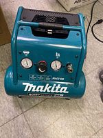 Front view showing pressure gauges, regulator knob, air outlet, and Makita branding on the air compressor.