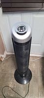 Front view of black and silver tower fan standing on tiled floor near white door with brown blinds above. Shows control buttons on top and vertical vented body.