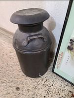 Vintage black metal milk can standing on tiled floor next to a framed picture, showing front side and lid with handle visible.