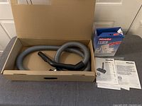 Vacuum hose with black handle attachments inside a cardboard box, placed beside Miele vacuum bags packaging and instruction manuals.