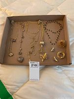 Full view of the collection in box showing five gold-toned necklaces laid flat along the top and right, with six pins grouped to the bottom right corner including butterfly, bee, knot with pearl, and starburst designs.