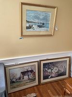 Wall-mounted rectangular framed print of a seashore scene with boats and figures, gold frame and cream mat, attributed to Monet. Below it, two more framed prints lean against a wall: one rural house scene, one floral still life.