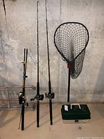 Three fishing rods with different reel types and landing net standing against concrete wall