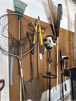 Photo showing various outdoor tools hung on pegboard including fishing net, rakes, shovel, string trimmer, and loppers