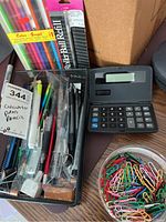 Photo showing pens, pencils, calculator, pen refills in black mesh organizer and brown box