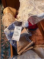 Assorted folded tablecloths, placemats, and napkins in various patterns including blue floral, beige fabric, and white crocheted lace.