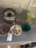 All items on display on a grey tablecloth: stainless steel tea kettle, vintage glass bowls, Vaseline glass hobnail fan vase, candy dish, ashtrays, and decorative plates and votive holders.